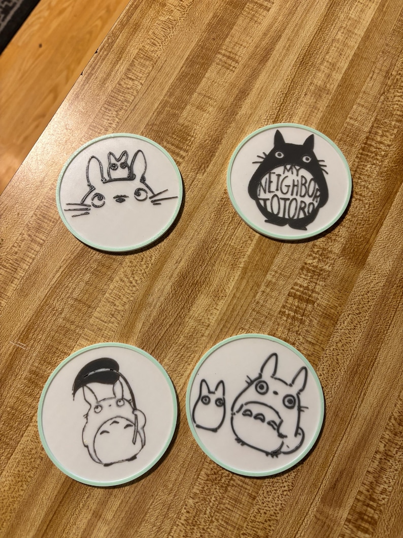 May include: Four circular white coasters with a light mint green border. Each coaster features a black line art illustration of characters from My Neighbor Totoro. One coaster includes the text My Neighbor Totoro in a bold black font.