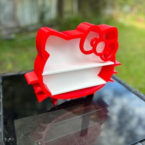 May include: A red, 3D-printed shelf shaped like a cartoon cat's head with a large bow. The shelf has two white shelves inside. The shelf is sitting on a black glass table, with a blurred green background.