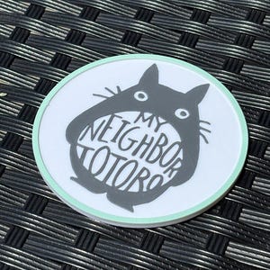 May include: A circular coaster with a white background and a light mint green border. The center features a dark gray silhouette of a creature with pointed ears and whiskers, containing the white text MY NEIGHBOR TOTORO inside its body.
