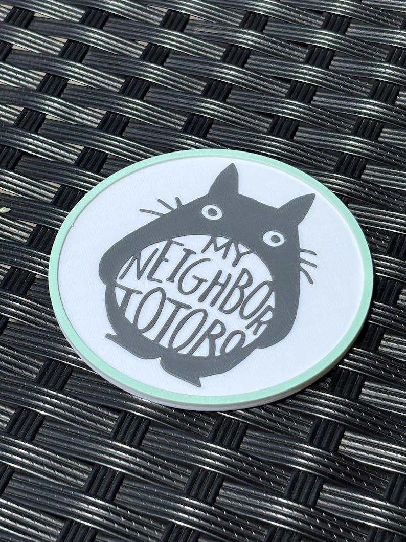 May include: A circular coaster with a white background and a light mint green border. The center features a dark gray silhouette of a creature with pointed ears and whiskers, containing the white text MY NEIGHBOR TOTORO inside its body.