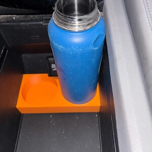 Puede incluir: Una botella de agua azul con tapa plateada se encuentra en un inserto naranja para portavasos. El inserto tiene un recorte circular para una taza y est&aacute; colocado en la consola central de un coche.