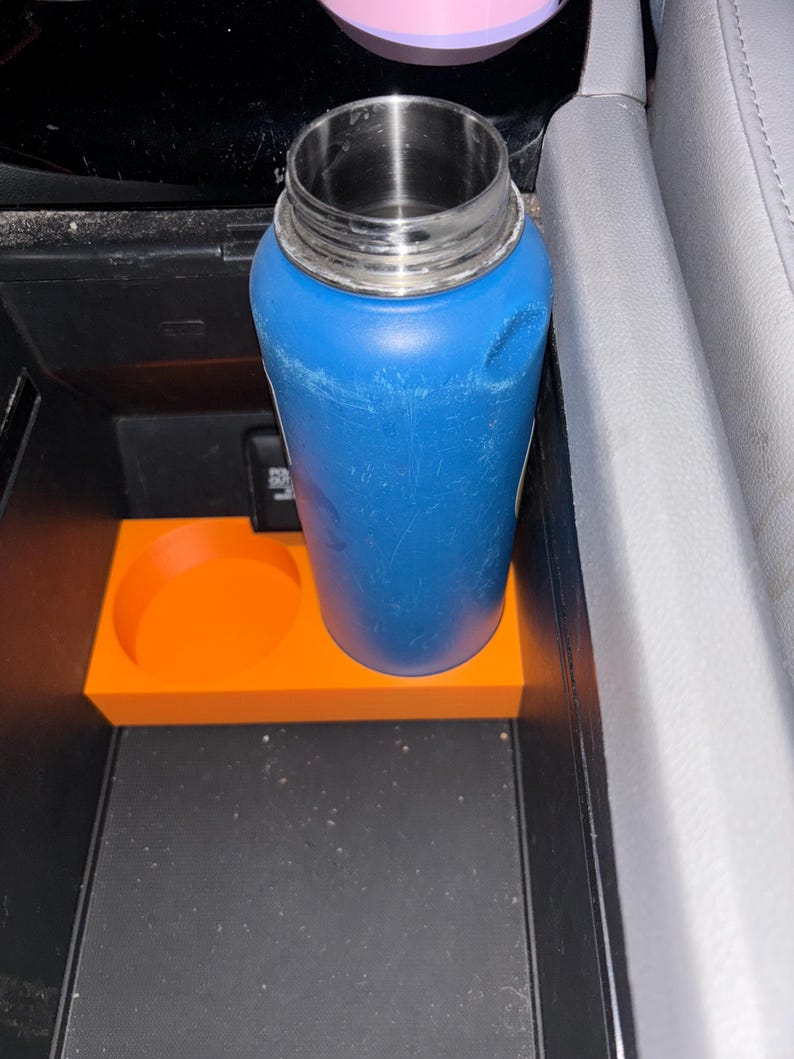 Puede incluir: Una botella de agua azul con tapa plateada se encuentra en un inserto naranja para portavasos. El inserto tiene un recorte circular para una taza y est&aacute; colocado en la consola central de un coche.