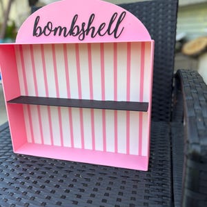 Puede incluir: Una estanter&iacute;a rosa con un estante negro y la palabra "bombshell" en escritura negra. El interior presenta rayas verticales rosas y blancas. La estanter&iacute;a est&aacute; sobre una silla de mimbre negra.