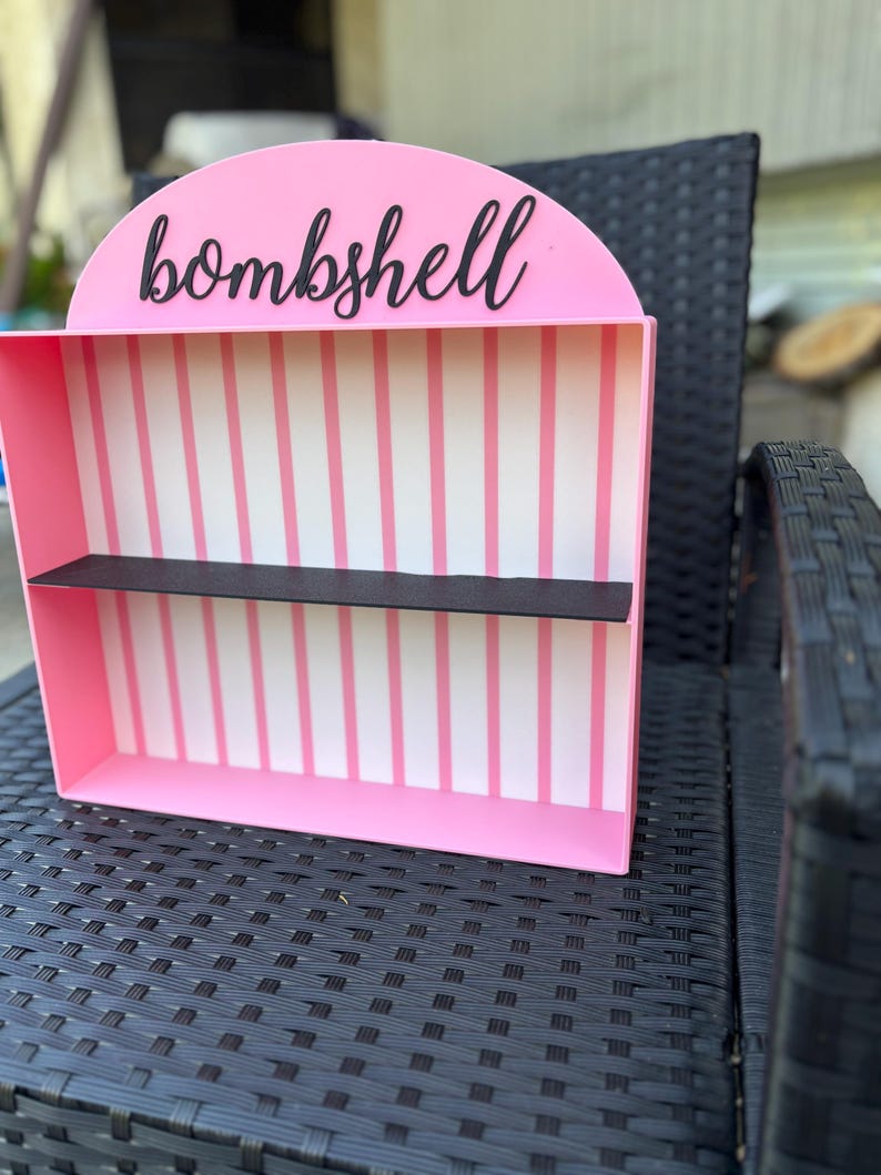 Puede incluir: Una estanter&iacute;a rosa con un estante negro y la palabra "bombshell" en escritura negra. El interior presenta rayas verticales rosas y blancas. La estanter&iacute;a est&aacute; sobre una silla de mimbre negra.