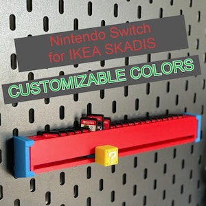 Puede incluir: Un soporte rojo y azul para cartuchos de juegos de Nintendo Switch para panel perforado IKEA SKÅDIS. El soporte tiene un bloque amarillo con un signo de interrogación. El texto dice "Nintendo Switch for IKEA SKADIS CUSTOMIZABLE COLORS."