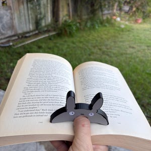 May include: A black and grey bunny-shaped bookmark with white eyes is placed on the pages of an open book. The bookmark is made of a glossy material. The book is open to pages 148 and 149.