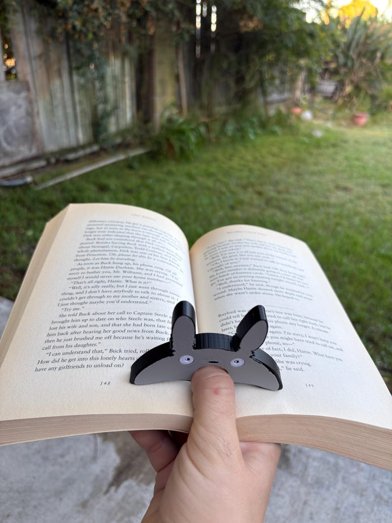 May include: A black and grey bunny-shaped bookmark with white eyes is placed on the pages of an open book. The bookmark is made of a glossy material. The book is open to pages 148 and 149.