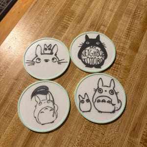 May include: A set of four circular white coasters with light green edges, each featuring black line art illustrations of characters from My Neighbor Totoro. One coaster displays the text My Neighbor Totoro inside the silhouette of the character.