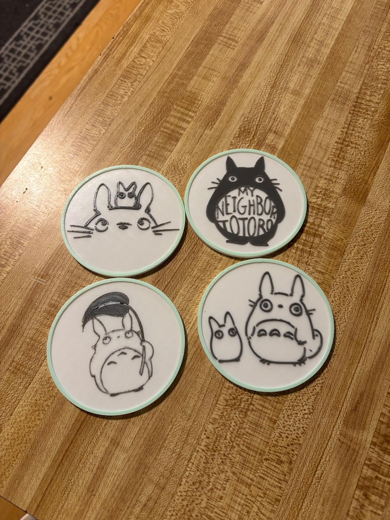 May include: A set of four circular white coasters with light green edges, each featuring black line art illustrations of characters from My Neighbor Totoro. One coaster displays the text My Neighbor Totoro inside the silhouette of the character.