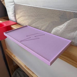 Puede incluir: Una estantería de mesita de noche de color lavanda con un diseño de rayo. La estantería rectangular tiene un borde elevado y está montada en un marco de cama de madera. Un portavasos rojo es visible en el lado izquierdo del marco.