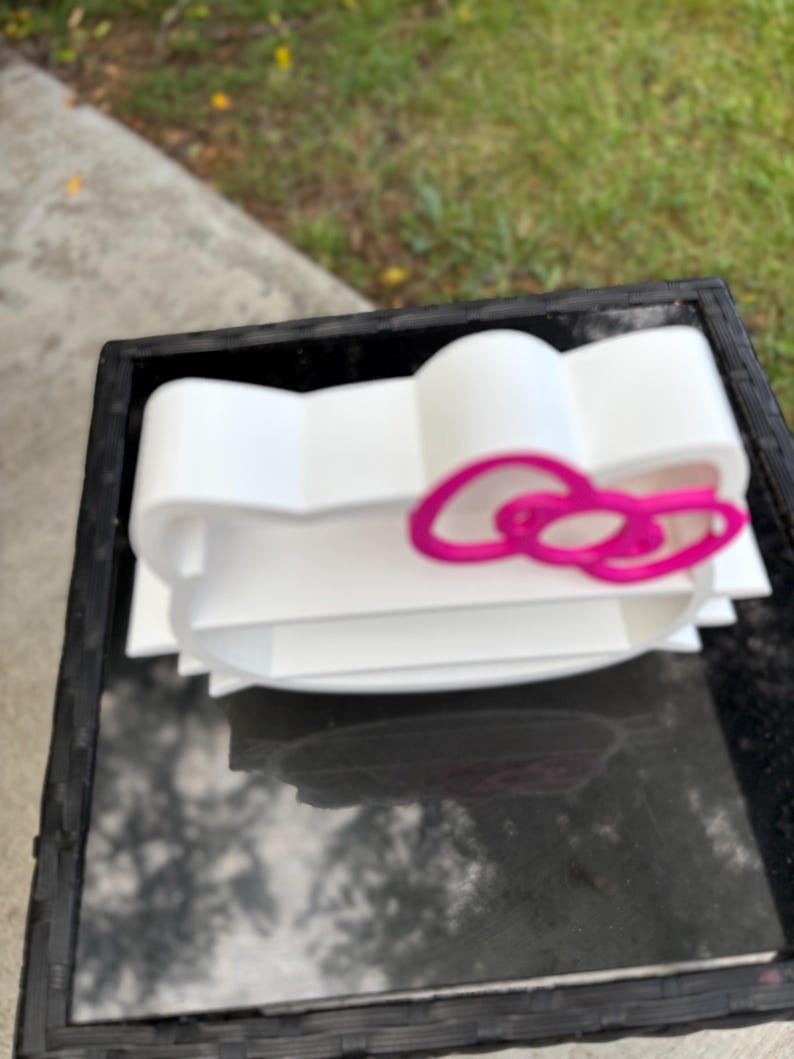 May include: A white, 3D-printed shelf in the shape of a cartoon cat's head, with a pink bow. The shelf has several levels for storage and is designed to be a decorative and functional item.