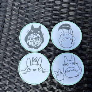 May include: Four circular coasters with a light green border, each featuring a black and white illustration of characters from My Neighbor Totoro. The top left coaster displays the text My Neighbor Totoro inside the silhouette of the large character.
