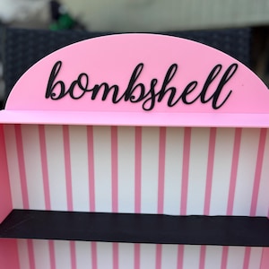 Puede incluir: Una estanter&iacute;a rosa con la palabra "bombshell" en escritura negra en la parte superior. El interior presenta paredes a rayas rosas y blancas y una estanter&iacute;a negra.