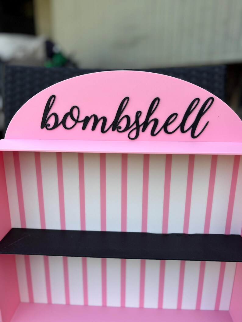 Puede incluir: Una estanter&iacute;a rosa con la palabra "bombshell" en escritura negra en la parte superior. El interior presenta paredes a rayas rosas y blancas y una estanter&iacute;a negra.