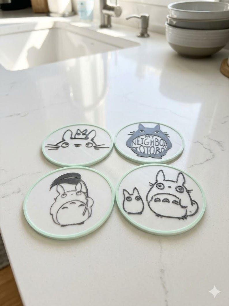 May include: Four circular white coasters with light green rims, each featuring black line art illustrations of characters from My Neighbor Totoro. The designs include various poses of the creatures, with one coaster displaying the text My Neighbor Totoro.