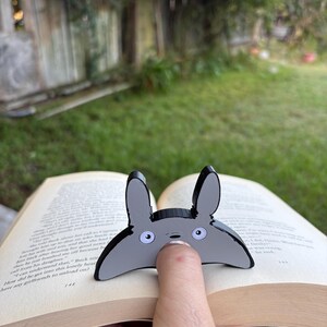 May include: A grey and black Totoro-shaped bookmark is placed on the pages of a book. The bookmark has large, round eyes and long ears. The book is open to page 148.