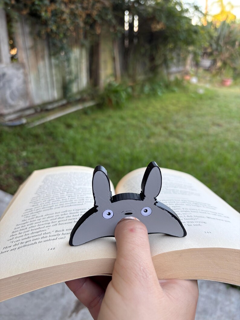 May include: A grey and black Totoro-shaped bookmark is placed on the pages of a book. The bookmark has large, round eyes and long ears. The book is open to page 148.