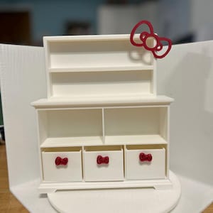 Könnte beinhalten: Weißes Miniatur-Puppenhausmöbel mit drei Schubladen und einem oberen Regal. Die Schubladen und das obere Regal haben rote Schleifenförmige Griffe. Das obere Regal hat eine große rote Schleife oben.