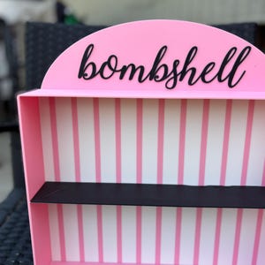 Puede incluir: Una estanter&iacute;a rosa con una balda negra y la palabra "bombshell" en escritura negra. El interior presenta rayas verticales rosas y blancas. La estanter&iacute;a tiene una parte superior redondeada.