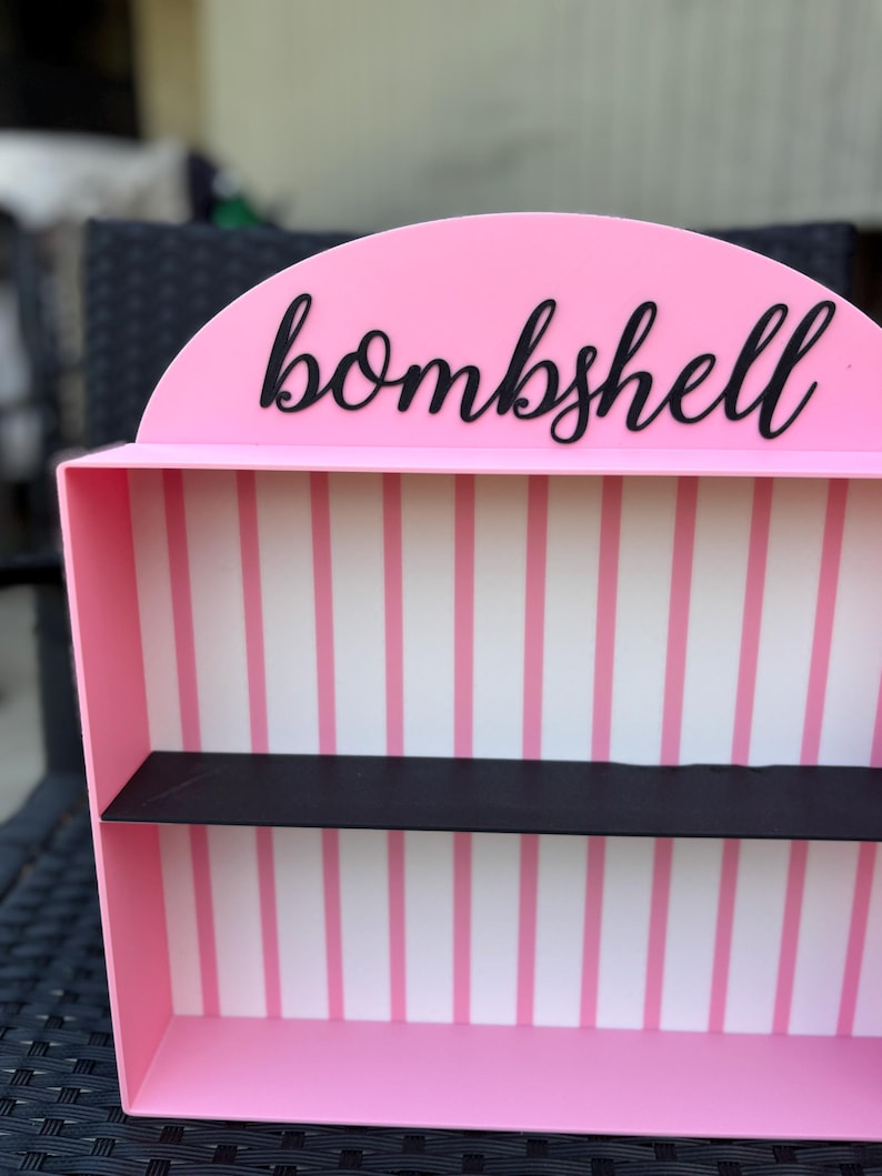 Puede incluir: Una estanter&iacute;a rosa con una balda negra y la palabra "bombshell" en escritura negra. El interior presenta rayas verticales rosas y blancas. La estanter&iacute;a tiene una parte superior redondeada.