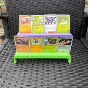 May include: A green and purple tiered display stand holding eight trading cards. The cards feature various colorful illustrations and text. The stand is on a black woven surface.