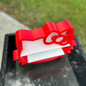 May include: A red, 3D-printed bow-shaped desk organiser with a white interior. The organiser is designed to hold papers or small items. The bow has a classic design and is placed on a glass surface.