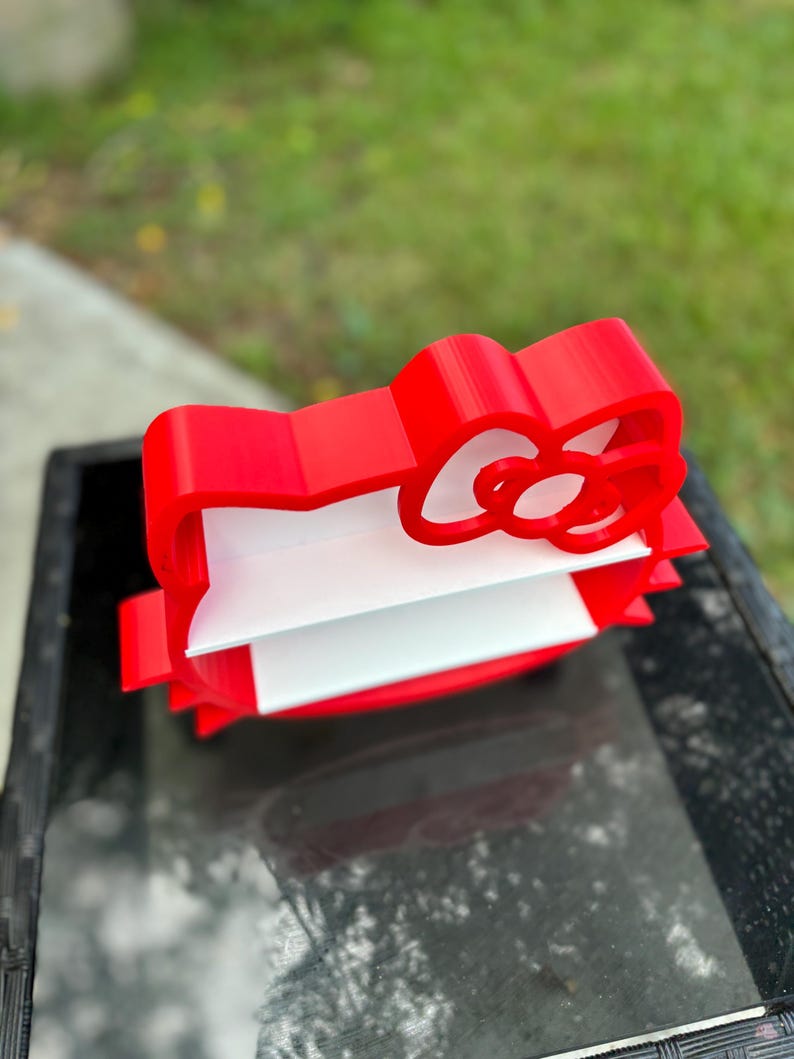 May include: A red, 3D-printed bow-shaped desk organiser with a white interior. The organiser is designed to hold papers or small items. The bow has a classic design and is placed on a glass surface.