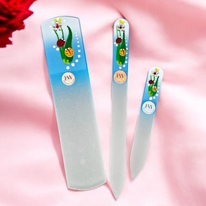 Puede incluir: Tres limas de uñas de vidrio con un diseño degradado en azul y blanco. Cada lima presenta un diseño floral con mariquitas y el texto "JW".