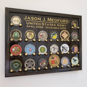 May include: A framed display of military challenge coins. The dark wood frame holds multiple gold-colored coins, each with unique designs and text. The display includes the name "JASON J. MEDFORD" and "UNITED STATES ARMY".