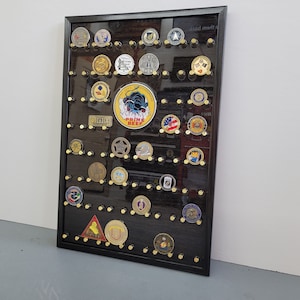 May include: A black framed shadow box with a black background and gold push pins. The shadow box is filled with various military challenge coins and patches. The coins and patches are arranged in rows and columns. The shadow box is hanging on a white wall.
