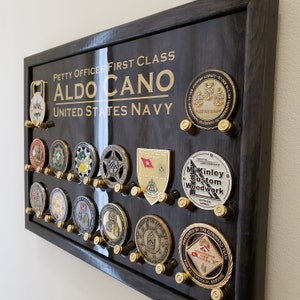May include: A black wooden shadow box with a gold inscription that reads "Petty Officer First Class Aldo Cano United States Navy". The shadow box is filled with military challenge coins and a bottle opener.