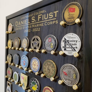 May include: A black wooden shadow box with gold lettering that reads "Lieutenant Colonel Daniel S. Fiust United States Marine Corps 2002-2022". The shadow box is filled with various military challenge coins and a few bullet casings.