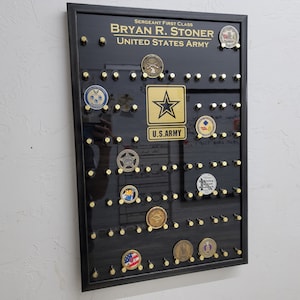May include: A black framed shadow box with a black background and gold accents. The shadow box has a gold star with the words "U.S. ARMY" underneath. The shadow box also has a gold coin with the words "Sergeant First Class Bryan R. Stoner United States Army".