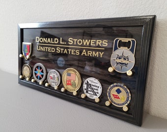 以下が含まれることがあります： 黒い木製の影箱に、金色の文字で「DONALD L. STOWERS UNITED STATES ARMY」と刻まれています。影箱には、栓抜きと、いくつかの軍隊のチャレンジコインが飾られています。
