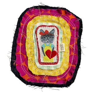 May include: A layered fabric patch featuring a central illustration of a grey cat wearing a red bow and yellow overalls, holding a red felt heart. The design includes concentric borders of yellow floral, light green, and pink polka dot fabric.