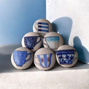 Op de afbeelding: Zeven ronde stoffen knopen, elk met een ander blauw en wit theekopje ontwerp. De illustraties van de theekopjes variëren in stijl, waaronder strepen, stippen en bloempatronen. De knopen zijn in een piramidevorm gerangschikt tegen een lichtblauwe achtergrond.