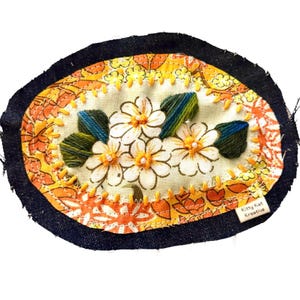 May include: An oval fabric patch featuring a central cream-coloured panel with embroidered white flowers, yellow centres, and green leaves. It is bordered by orange floral fabric and a dark denim edge, with a small tag reading Kitty Kat Kreative.