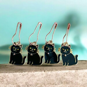 May include: Four black cat stitch markers with gold detailing and safety pin fastenings. Each cat has yellow eyes and a small pink collar. The markers are displayed on a grey surface against a teal backdrop.