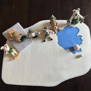 May include: A white felt play mat with a blue felt lake and a white felt border. There are several small felt animal toys on the mat, including a bear, a squirrel, a rabbit, and a fox. There are also two white felt balls on the mat.