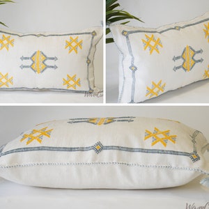 May include: White throw pillow with a geometric pattern in yellow and blue embroidery. The pillow has a fringe trim.