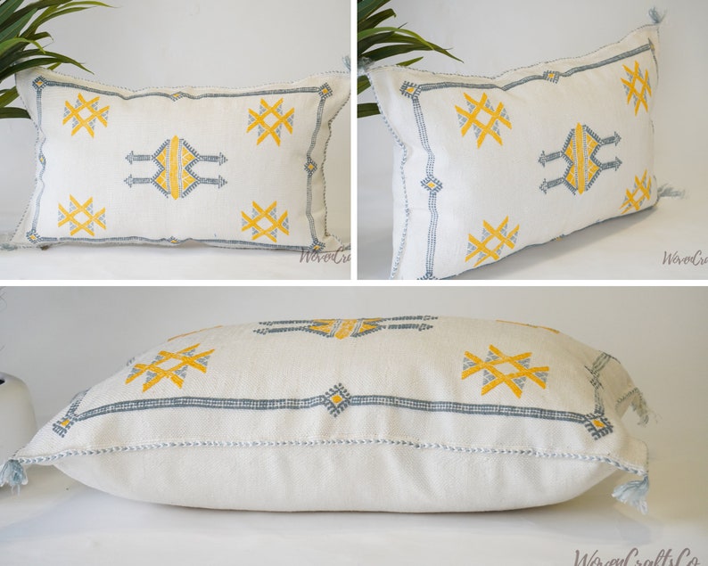 May include: White throw pillow with a geometric pattern in yellow and blue embroidery. The pillow has a fringe trim.