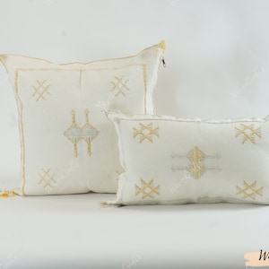 May include: Two white decorative throw pillows with embroidered geometric patterns in gold and silver. The square pillow has a zipper closure and gold tassels. The rectangular pillow has gold and silver tassels. The pillows are on a white background.