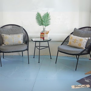 May include: Two black wicker chairs with gray cushions and patterned throw pillows, a small black metal table with a glass top, and a vase with a green leafy plant. The chairs and table are on a light blue floor.