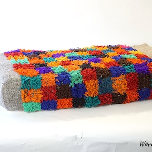 May include: A colorful, square-shaped pouf with a patchwork design. The pouf is made of a soft, fluffy material and has a gray fabric border.