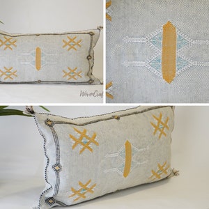 May include: A white and gray rectangular pillow with a geometric pattern in yellow and blue thread. The pillow has a fringe on the edges.
