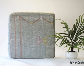 May include: A gray square ottoman with a woven pattern in pink, yellow, and red. The ottoman is sitting on a white surface in front of a green plant.