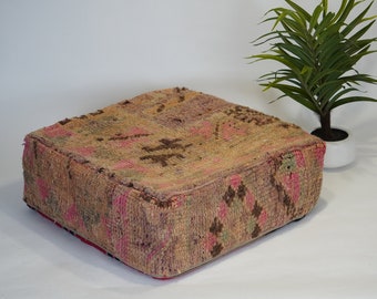 May include: A square, woven floor cushion with a pink, brown, and beige pattern. The cushion is made of a soft, textured fabric and has a slightly distressed look.