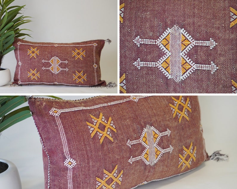 May include: A burgundy throw pillow with a geometric pattern in white and yellow thread. The pillow has a fringe border and is made of a soft, woven fabric.