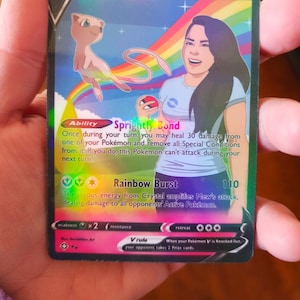 Puede incluir: Una carta de comercio de Pokemon holográfica con Crystal & Mew. La carta tiene un fondo de arcoíris y presenta una ilustración de dibujos animados de un Mew rosa y una mujer con cabello largo y castaño. El texto de la carta dice 'Sprightly Bond' y 'Rainbow Burst'.