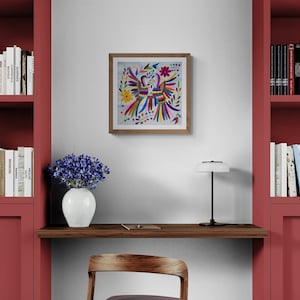 May include: A square framed wall art piece featuring a colorful embroidered-style design of two birds surrounded by flowers and leaves, mounted on a white wall above a wooden desk with a white vase of blue flowers and a black desk lamp.